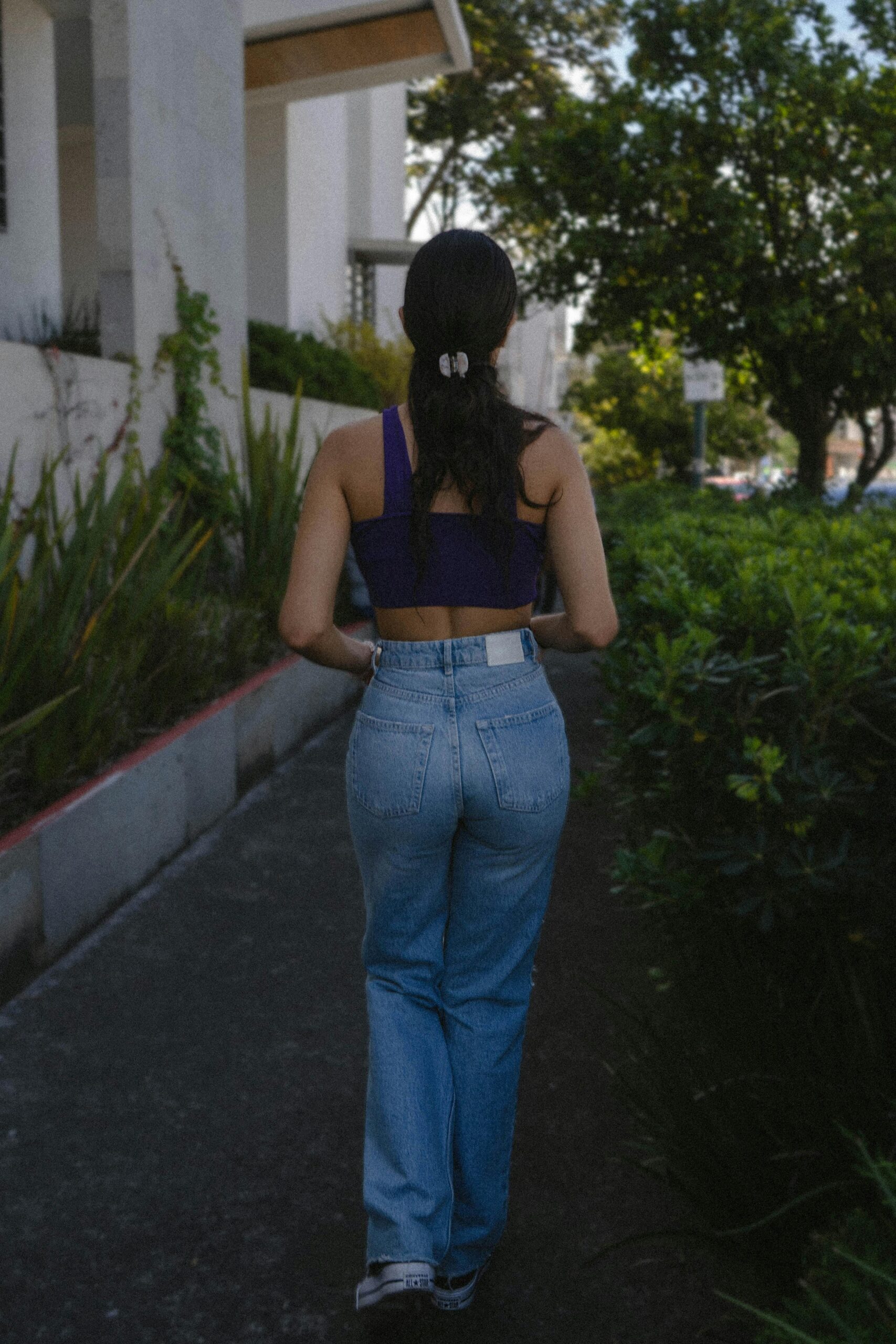 Back view of a woman walking on a verdant city sidewalk during daylight.