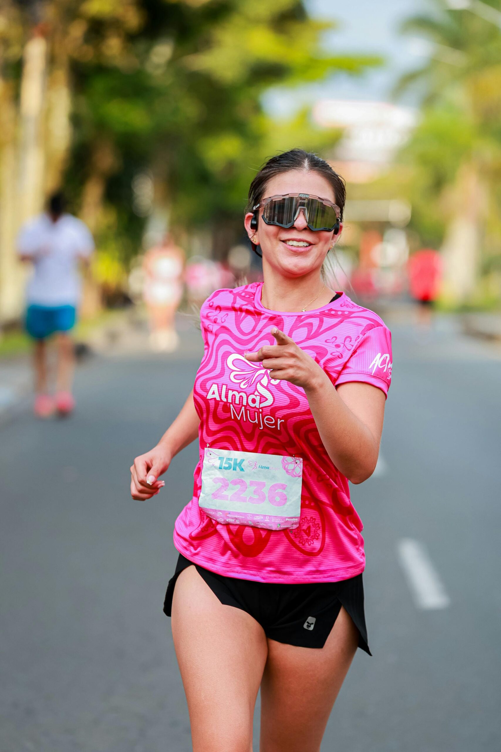 A smiling woman running in a 15K race, wearing a pink 'Alma Mujer' shirt and sunglasses.