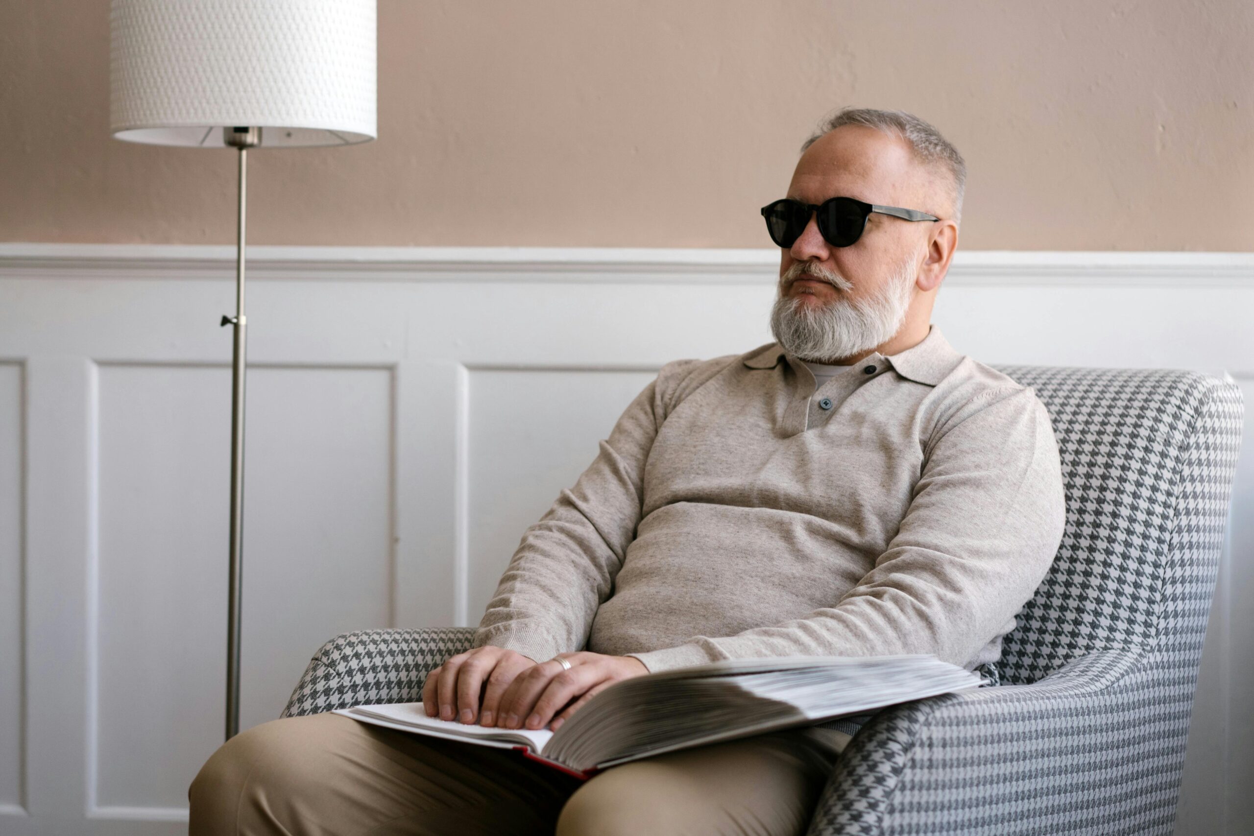 Senior man with sunglasses reading a braille book in a cozy modern interior.